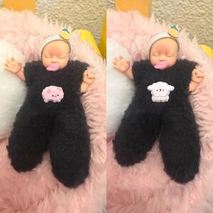 May include: A doll wearing a black fuzzy jumpsuit with a pink pig or sheep applique. The doll has a white headband with a yellow and green fruit decoration. The doll is lying on a pink and white fluffy surface.
