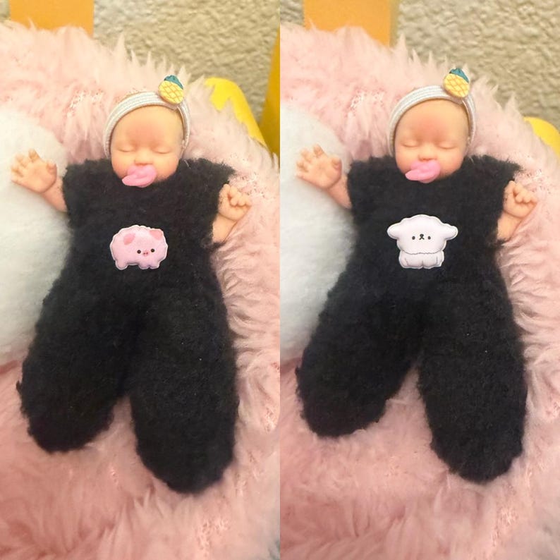 May include: A doll wearing a black fuzzy jumpsuit with a pink pig or sheep applique. The doll has a white headband with a yellow and green fruit decoration. The doll is lying on a pink and white fluffy surface.