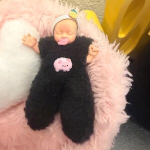 May include: A doll dressed in a black fuzzy jumpsuit with a pink pig patch. The doll has a headband with a pineapple and a pacifier. It is lying on a pink fluffy surface.