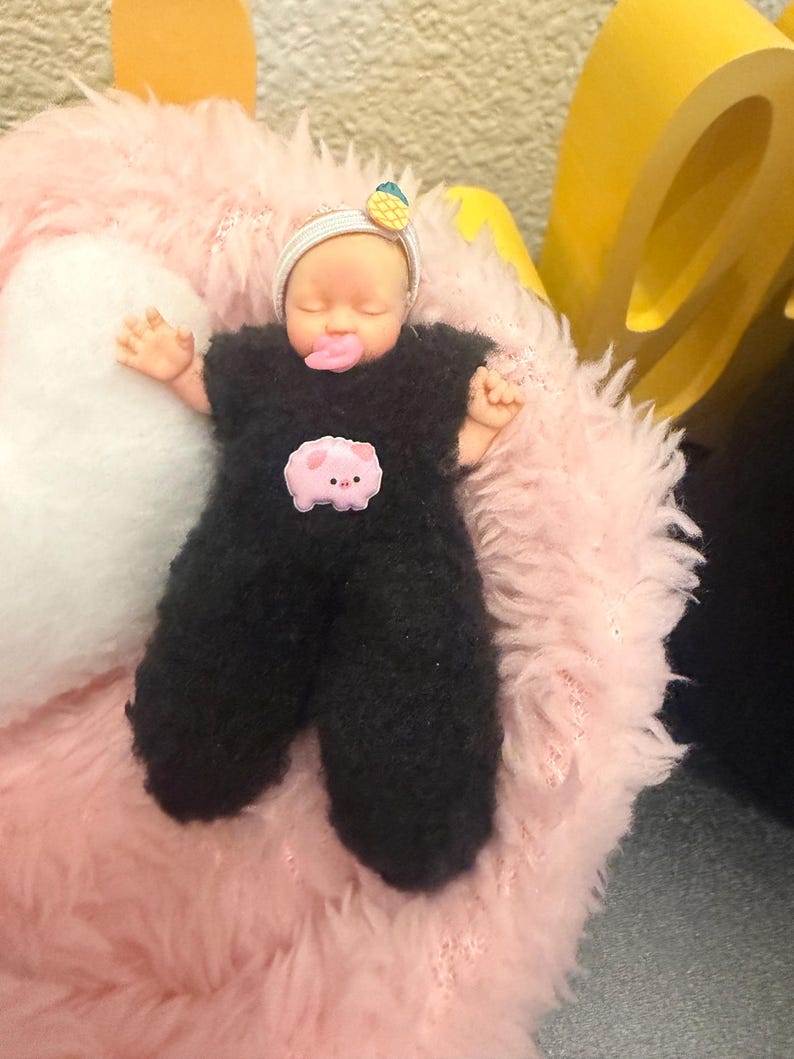 May include: A doll dressed in a black fuzzy jumpsuit with a pink pig patch. The doll has a headband with a pineapple and a pacifier. It is lying on a pink fluffy surface.