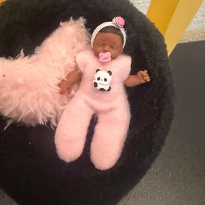 May include: A miniature baby doll with dark skin, wearing a pink onesie with a panda bear graphic. The doll has a pacifier and a white headband with a pink flower. It rests on a black surface with a pink fluffy blanket.