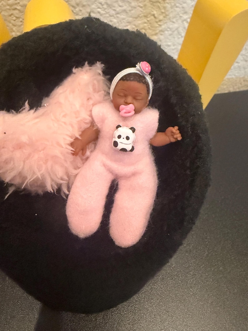 May include: A miniature baby doll with dark skin, wearing a pink onesie with a panda bear graphic. The doll has a pacifier and a white headband with a pink flower. It rests on a black surface with a pink fluffy blanket.