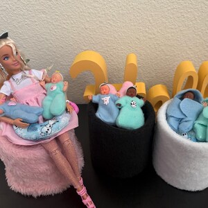 May include: A Barbie doll in a pink dress and accessories, holding three baby dolls. Two black felt containers and one white felt container each hold two baby dolls wrapped in blue and teal blankets. A decorative yellow number is in the background.
