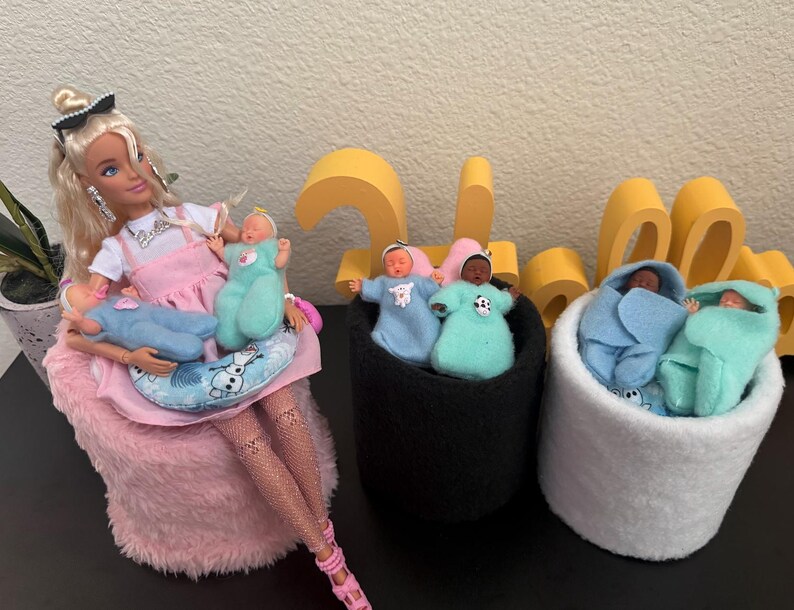 May include: A Barbie doll in a pink dress and accessories, holding three baby dolls. Two black felt containers and one white felt container each hold two baby dolls wrapped in blue and teal blankets. A decorative yellow number is in the background.