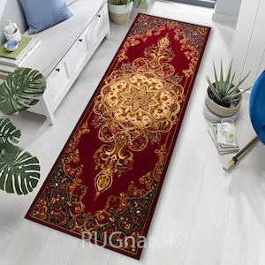 Może przedstawiać: Prostokątny dywanik z głębokim czerwonym tłem i ozdobnym, symetrycznym wzorem. Centralny motyw to duży, kremowy wzór kwiatowy ze złotymi i brązowymi akcentami. Krawędzie dywanu zdobią misterne detale.