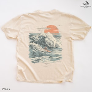 Puede incluir: Camiseta de color marfil con un gráfico de un surfista surfeando una ola al atardecer. El diseño incluye un gran sol naranja, olas rompiendo y el texto "Chasing Waves". La camiseta es de un material suave.
