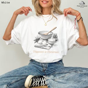 May include: White t-shirt with a watercolor illustration of baking ingredients, including a bowl, eggs, and flour. The text "Happiness is Homemade" is printed below the illustration. The shirt is a casual, comfortable style.