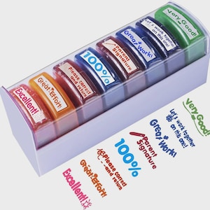 May include: A set of six colorful self-inking teacher stamps in a light purple plastic storage tray. The stamps feature phrases including Excellent!, Great Effort!, Please correct and return, 100%, Parent Signature, Great Work!, and Let's work together on this one!.