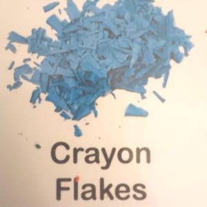 May include: A close-up of a pile of blue crayon flakes. The flakes are scattered on a white surface. The words "Crayon Flakes" are printed in black below the flakes.