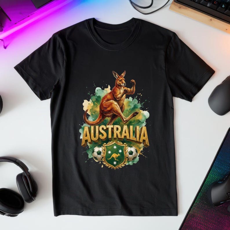 May include: Black t-shirt featuring a watercolor illustration of a kangaroo flexing its muscles. The design includes the word "AUSTRALIA" in gold lettering, a soccer ball, and a green and gold crest.