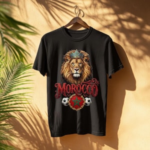 May include: Black t-shirt featuring a lion wearing a crown, with the word "MOROCCO" in red script. Below the lion is a red and green emblem with a star, flanked by soccer balls. The shirt is hanging on a wooden hanger.
