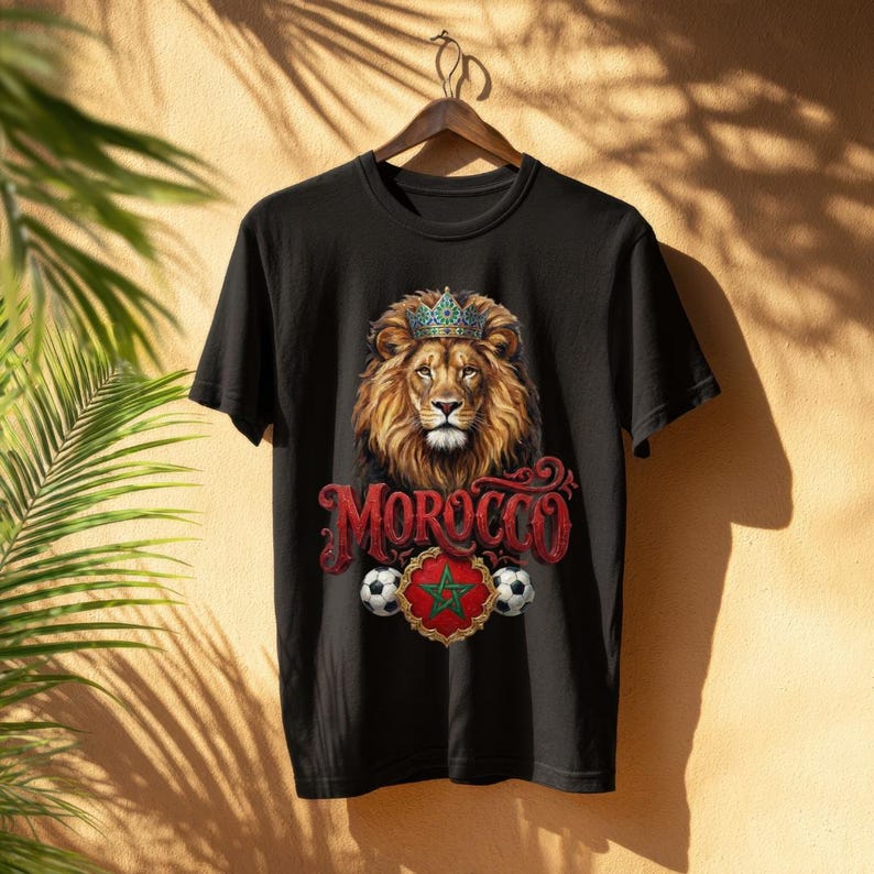 May include: Black t-shirt featuring a lion wearing a crown, with the word "MOROCCO" in red script. Below the lion is a red and green emblem with a star, flanked by soccer balls. The shirt is hanging on a wooden hanger.