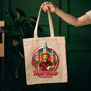 May include: Beige tote bag featuring a lion wearing a red jersey and holding a soccer ball, with the text "Morocco WORLD CUP 2026". The design includes a circular emblem with Moroccan imagery.