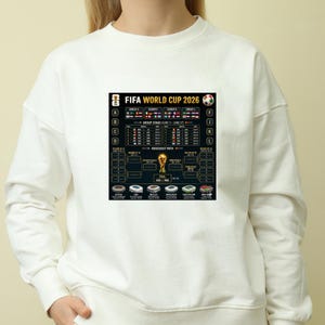 May include: White sweatshirt featuring a graphic of the FIFA World Cup 2026 schedule. The design includes team flags, group stage details, and a knockout path. The text "FIFA WORLD CUP 2026" is prominently displayed.