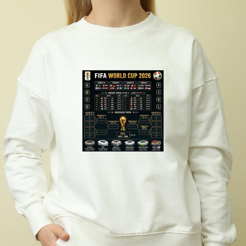 May include: White sweatshirt featuring a graphic of the FIFA World Cup 2026 schedule. The design includes team flags, group stage details, and a knockout path. The text "FIFA WORLD CUP 2026" is prominently displayed.