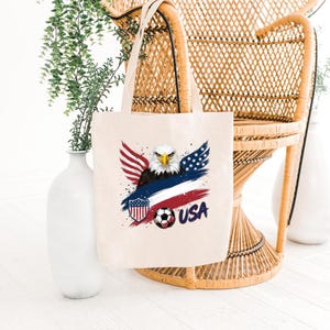 May include: A cream-colored tote bag featuring a patriotic design. The graphic includes a bald eagle with red, white, and blue wings, a soccer ball, and the word "USA". The bag is made of a sturdy fabric and has two handles.