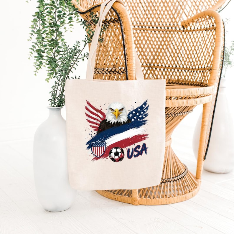 May include: A cream-colored tote bag featuring a patriotic design. The graphic includes a bald eagle with red, white, and blue wings, a soccer ball, and the word "USA". The bag is made of a sturdy fabric and has two handles.