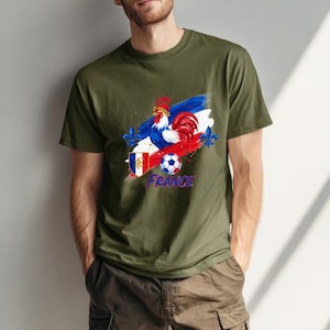 May include: Olive green t-shirt featuring a graphic of a rooster, a soccer ball, and the word "France" in blue. The design incorporates the colors of the French flag: red, white, and blue. The shirt has a crew neck and short sleeves.
