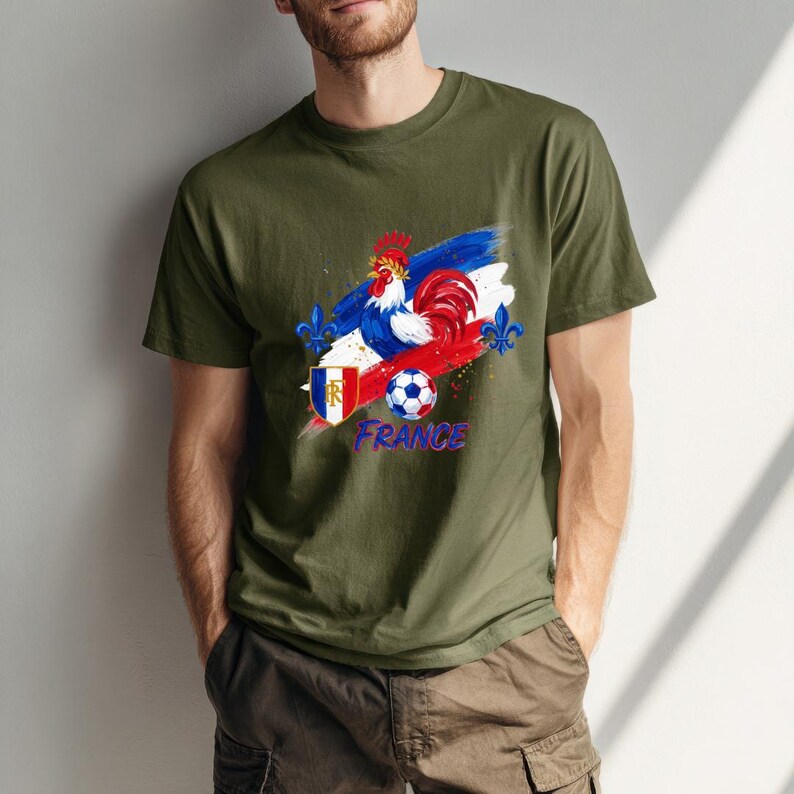 May include: Olive green t-shirt featuring a graphic of a rooster, a soccer ball, and the word "France" in blue. The design incorporates the colors of the French flag: red, white, and blue. The shirt has a crew neck and short sleeves.