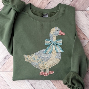 May include: An olive green sweatshirt featuring a goose applique. The goose has a blue and white patterned body, a red and white checkered beak and feet, and a blue and white plaid bow. The sleeves have a gathered cuff.