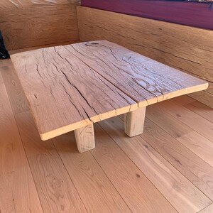 May include: A rustic, square wooden coffee table with a light brown finish. The tabletop is made of two planks with visible wood grain and cracks. It rests on two short, square legs, showcasing a natural, unfinished aesthetic.