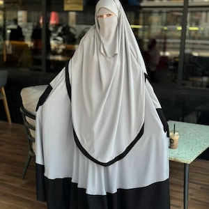 Könnte beinhalten: Ein hellgraues Abaya- und Niqab-Set mit schwarzem Besatz. Die Abaya ist ein langes, fließendes Kleidungsstück mit weitem Rock. Der Niqab bedeckt das Gesicht und lässt nur die Augen frei. Das Outfit wird mit schwarzen Handschuhen getragen.