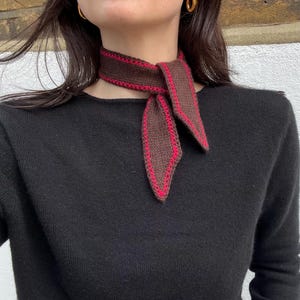 Puede incluir: Bufanda de punto marrón y roja atada al cuello. La bufanda tiene una base marrón oscuro con un borde rojo. Se lleva sobre un jersey negro. Un accesorio de moda.