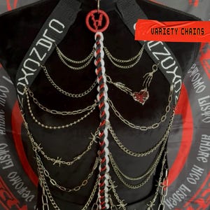 May include: A black harness featuring a choker collar, white text on straps, and a central braided cord in red, black, and white. The design includes multiple silver-toned metal chains, barbed wire accents, and a skeletal hand holding a red heart charm.
