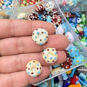 Könnte beinhalten: Nahaufnahme von handgefertigten Glasperlen in verschiedenen Formen und Farben. Drei donut-förmige Perlen mit weißem Zuckerguss, braunen Zentren und blauen und braunen Streuseln stehen im Fokus. Weitere Perlen sind im Hintergrund zu sehen.