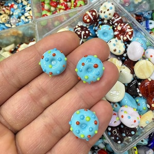 Könnte beinhalten: Nahaufnahme von drei hellblauen, donut-förmigen Glasperlen mit bunten Punktakzenten. Die Perlen sind von anderen bunten Perlen in verschiedenen Formen und Größen umgeben. Diese Perlen sind ideal für die Schmuckherstellung und das Basteln.