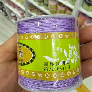 May include: A spool of purple thread is encased in clear plastic. The label is green with white floral designs and Chinese characters. The thread is tightly wound, and the spool is held in a hand.