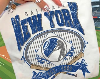 Op de afbeelding: Wit T-shirt met een blauwe vintage graphic met de tekst "BASEBALL NEW YORK" en een silhouet van een honkbalspeler. Het ontwerp bevat gekruiste honkbalknuppels en de tekst "EST. 1973".