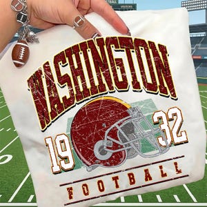 May include: A white t-shirt featuring a distressed graphic with the text WASHINGTON in large maroon letters, the number 1932, and a maroon football helmet. The design includes the word FOOTBALL at the bottom, all set against a green field background.