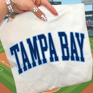 May include: White t-shirt with the text "TAMPA BAY" in dark blue block letters with a light blue outline. The shirt is folded, and the background features a baseball field.