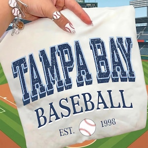 May include: Cream-colored t-shirt featuring the text "TAMPA BAY BASEBALL" in navy blue with a light blue outline. A baseball graphic and the text "EST. 1998" are also present. The shirt is folded.