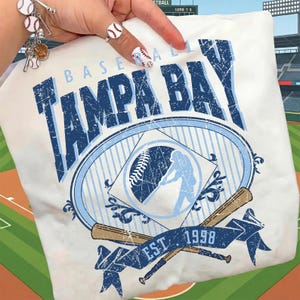 May include: White t-shirt featuring a distressed design with the text "TAMPA BAY BASEBALL" in blue. The design includes a baseball, a batter silhouette, crossed bats, and the text "EST 1998".