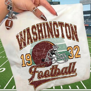 May include: A white sweatshirt featuring a distressed graphic with the text WASHINGTON 1932 Football and a maroon football helmet. The design includes a green striped background element, with maroon and gold lettering throughout the print.