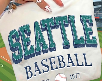 May include: A white t-shirt with the words "SEATTLE BASEBALL" in teal and navy blue, with a baseball graphic and "EST. 1977". The shirt is displayed on a baseball field background.