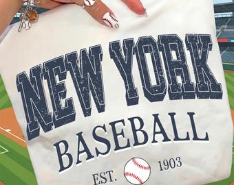 Op de afbeelding: Wit t-shirt met de tekst "NEW YORK BASEBALL" in marineblauw, een honkbalafbeelding en het jaar "1903". Het shirt wordt gepresenteerd tegen een honkbalveldachtergrond.