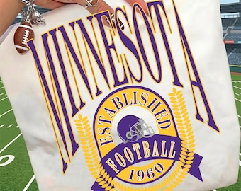 May include: A white t-shirt featuring the purple and yellow text MINNESOTA, with a circular emblem containing the words ESTABLISHED FOOTBALL 1960, a football helmet graphic, and wheat stalks. The design is printed in a collegiate athletic style.