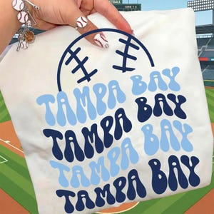 May include: White t-shirt featuring the text "TAMPA BAY" in light and dark blue, with a baseball graphic. The shirt is held up by a hand with baseball-themed jewelry and nail art, set against a baseball field backdrop.
