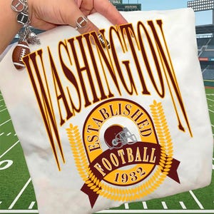 May include: A white t-shirt featuring the word WASHINGTON in large burgundy letters. Below, a circular emblem displays the text ESTABLISHED FOOTBALL 1932, a football helmet graphic, and yellow wheat stalks. The design is printed in burgundy and yellow.