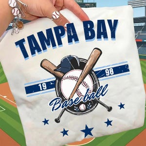 May include: White t-shirt featuring the text "TAMPA BAY" in navy blue, with a baseball graphic. The graphic includes crossed bats, a baseball, a baseball cap, and the word "Baseball". Blue stars are scattered around the graphic.