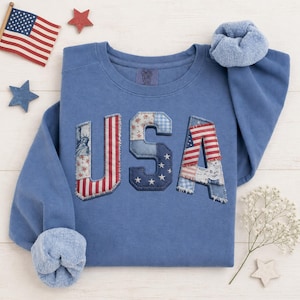 May include: A blue sweatshirt with the word "USA" appliqued on the front. The letters are made of various patriotic fabrics, including the American flag, stars, and floral patterns. The sleeves have a soft, fuzzy lining.