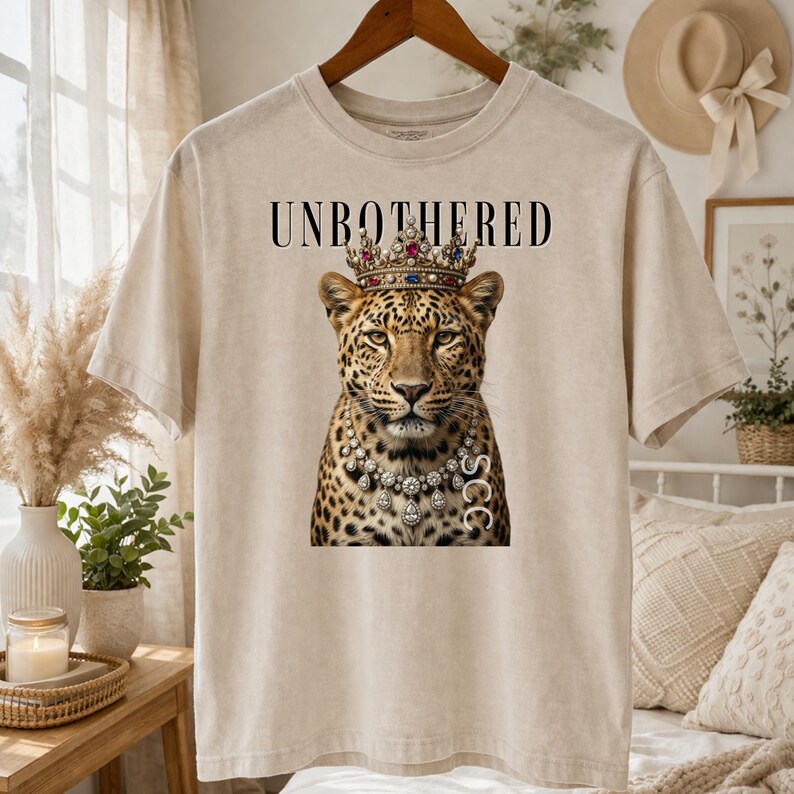 May include: Beige t-shirt featuring a leopard wearing a crown and a diamond necklace. The text "UNBOTHERED" is printed above the leopard's head. The leopard has a regal expression.