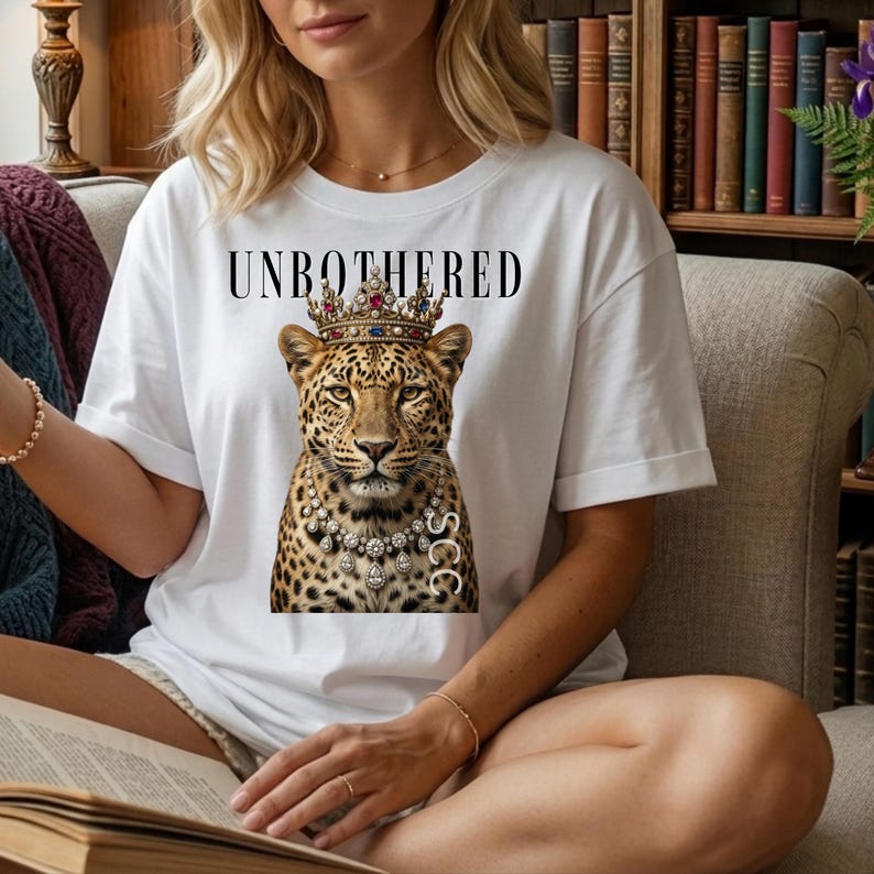 May include: White t-shirt featuring a leopard wearing a jeweled crown and necklace. The text "UNBOTHERED" is printed above the leopard. The leopard has a detailed spotted pattern. The letters "SCC" are printed on the necklace.