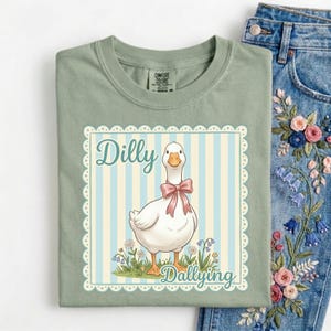 May include: Sage green t-shirt featuring a cartoon goose with a pink bow, standing in a field of flowers. The words "Dilly Dallying" are printed on the shirt in a decorative frame. The shirt is folded next to a pair of embroidered jeans.