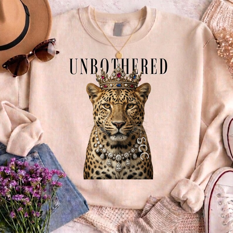 May include: A light pink sweatshirt with the word "UNBOTHERED" printed across the top. The sweatshirt features a leopard wearing a jeweled crown and necklace. Sunglasses and a hat are visible in the background.
