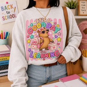 May include: A light gray crewneck sweatshirt featuring a graphic of a brown bear wearing sunglasses and a pink bow, carrying a backpack and lunchbox, with the text IT'S A GOOD DAY FOR LEARNING surrounded by colorful stars and hearts.