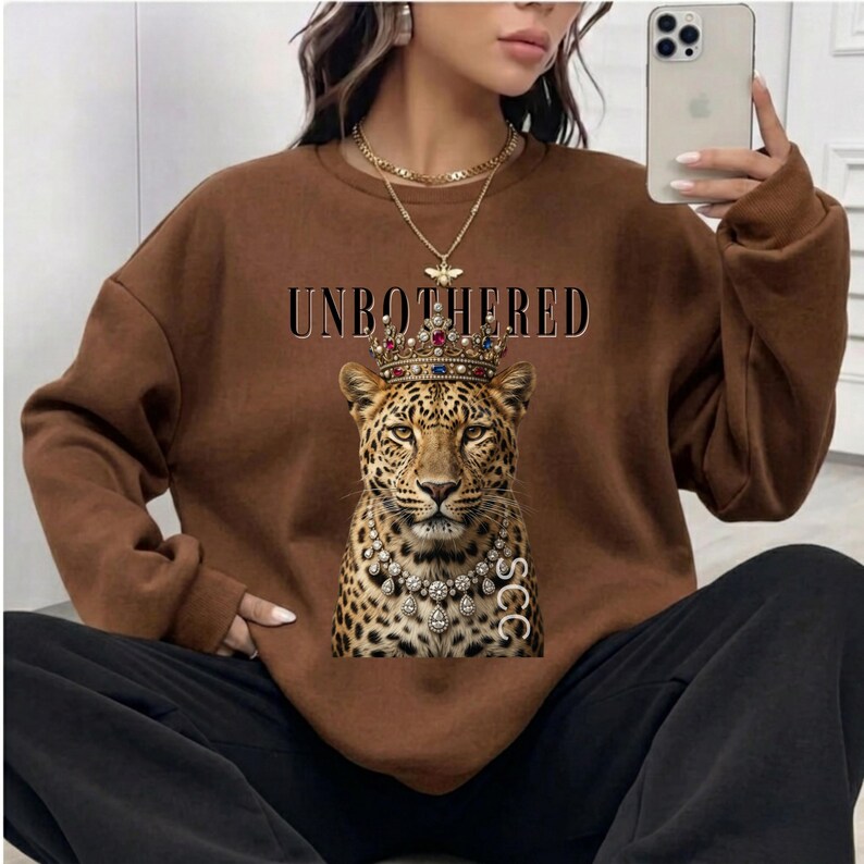 May include: Brown sweatshirt with the word "UNBOTHERED" above a leopard wearing a jeweled crown and necklace. The leopard graphic is centered on the front of the sweatshirt.